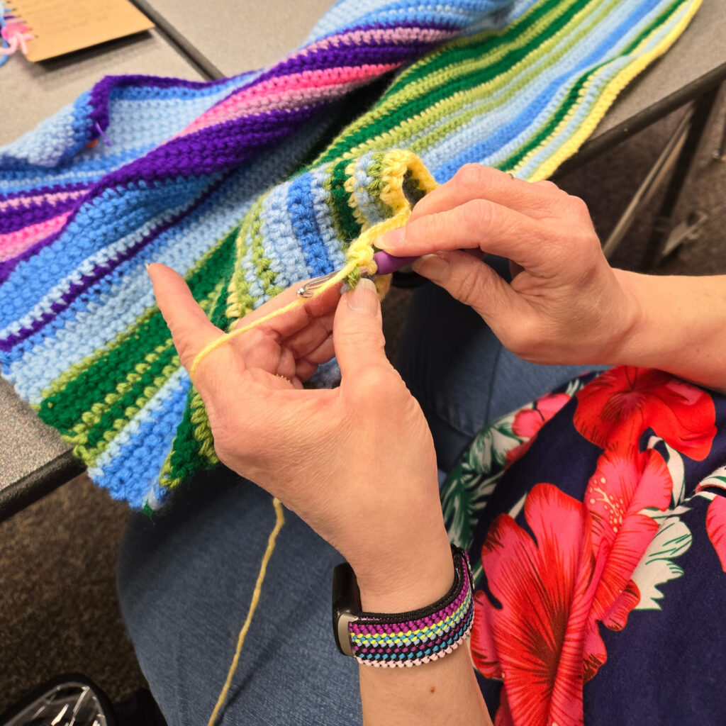 A person crocheting a colorful striped blanket with a purple crochet hook and yellow yarn.