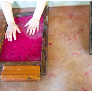 Two hands pressing pink pulp mixture in a wooden frame for paper making on a light brown surface.
