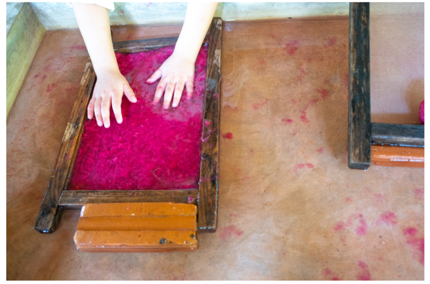 Two hands pressing pink pulp mixture in a wooden frame for paper making on a light brown surface.